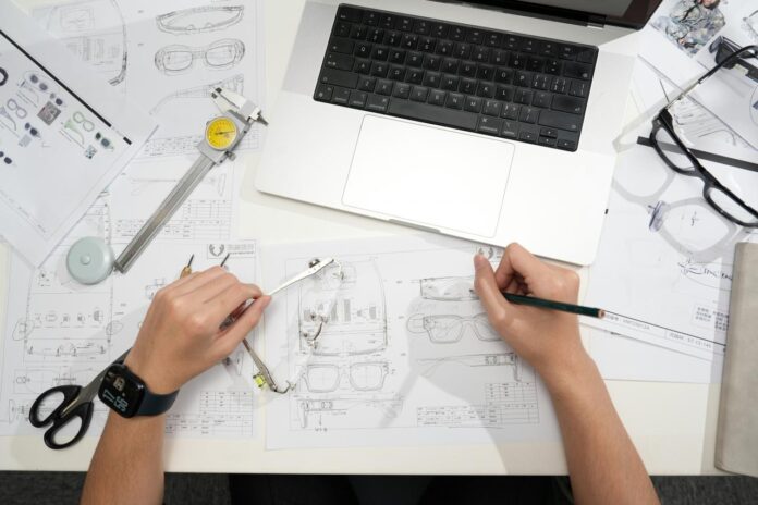 Desainer membuat sketsa kacamata dengan laptop dan alat.