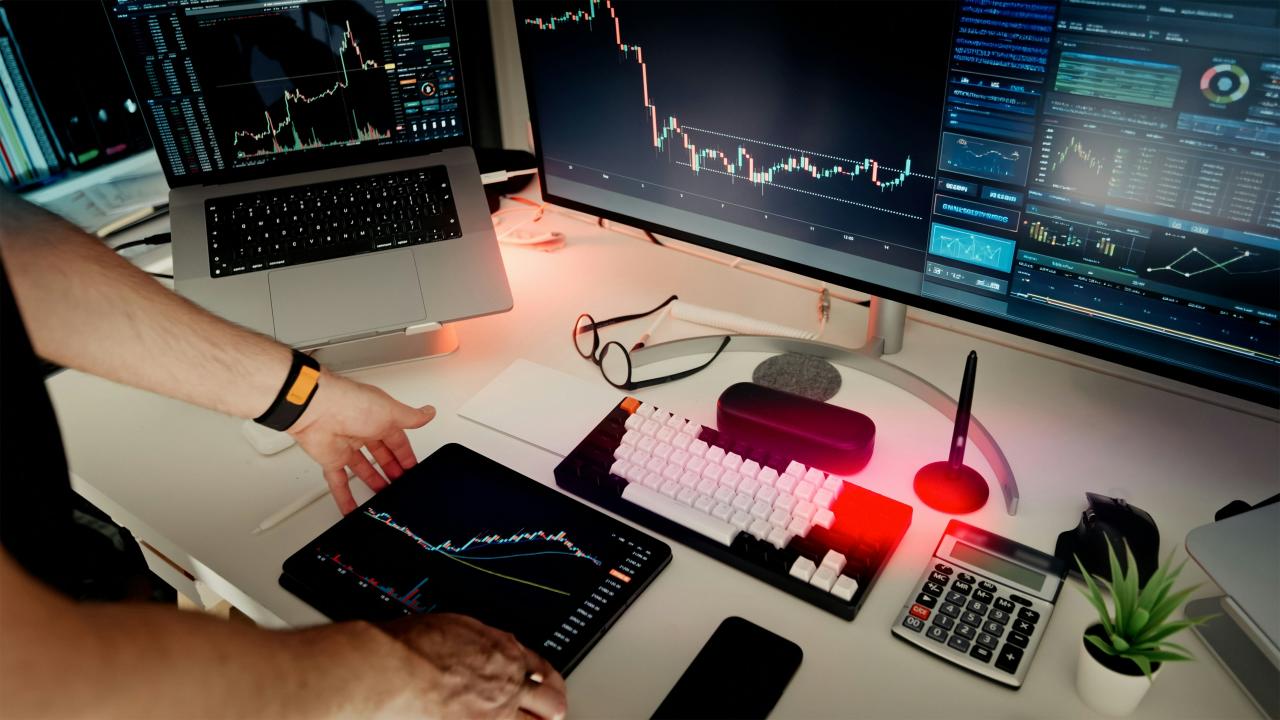 Seorang pria duduk di meja dengan dua monitor dan laptop